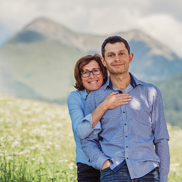 Die Gastgeber Kurt und Monika lächeln, mit den Dolomiten und einer blühenden Wiese von Maranza im Hintergrund.