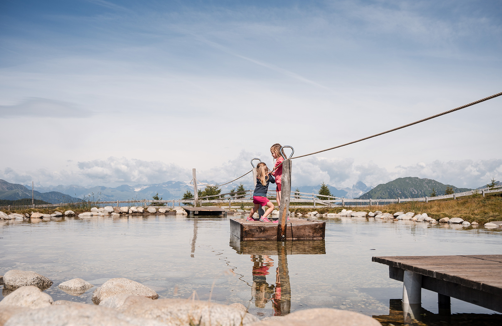 Zwei Mädchen spielen auf einem Floß in einem alpinen Teich, mit den Dolomiten im Hintergrund. Perfekt für Familien im Urlaub in Maranza.