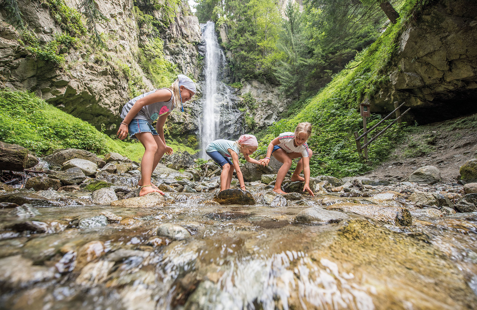 Drei Mädchen spielen glücklich in einem Bergbach mit einem Wasserfall im Hintergrund, umgeben von der Natur Südtirols.