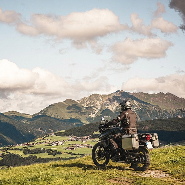 Motorradfahrer mit Reiseausrüstung bewundert das Panorama der Dolomiten und des Pustertals, mit einem Alpendorf darunter.