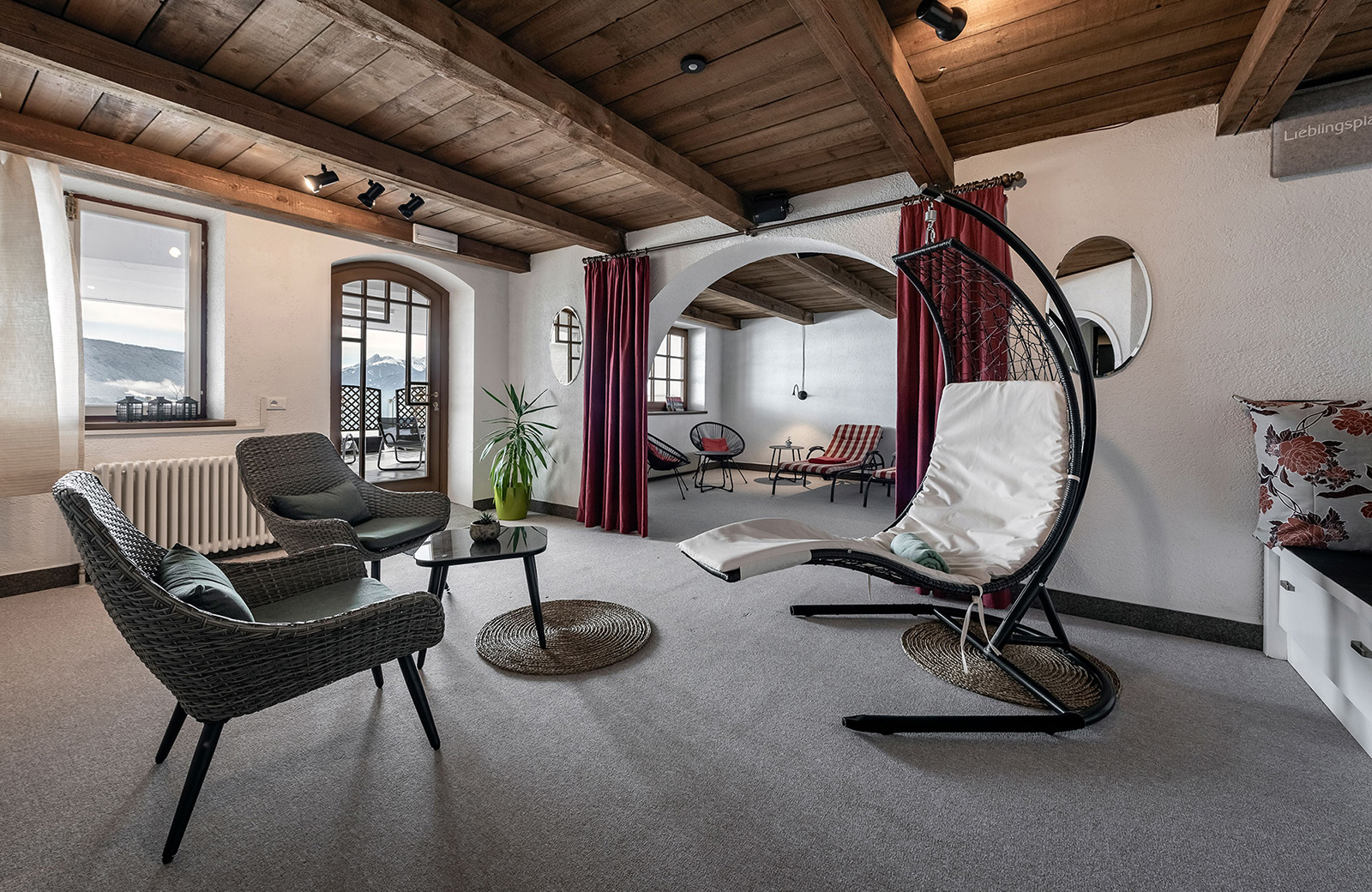 Spacious relaxation room with a wooden ceiling, wicker armchairs, a suspended chaise longue, and a view of the Dolomites.
