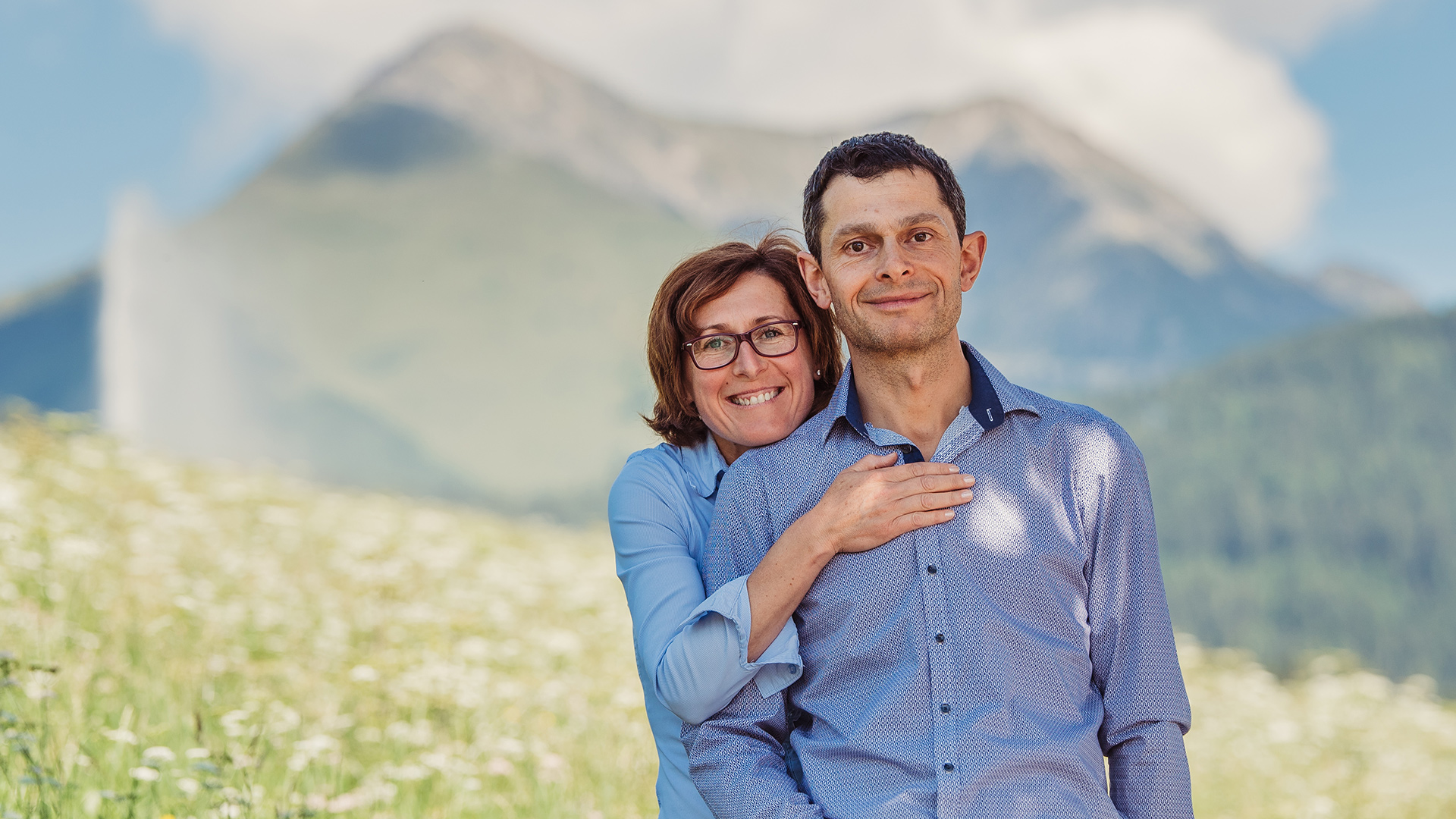 Kurt und Monika, die lächelnden Betreiber der Pension Wiesenrain, mit den Dolomiten und einer Blumenwiese.