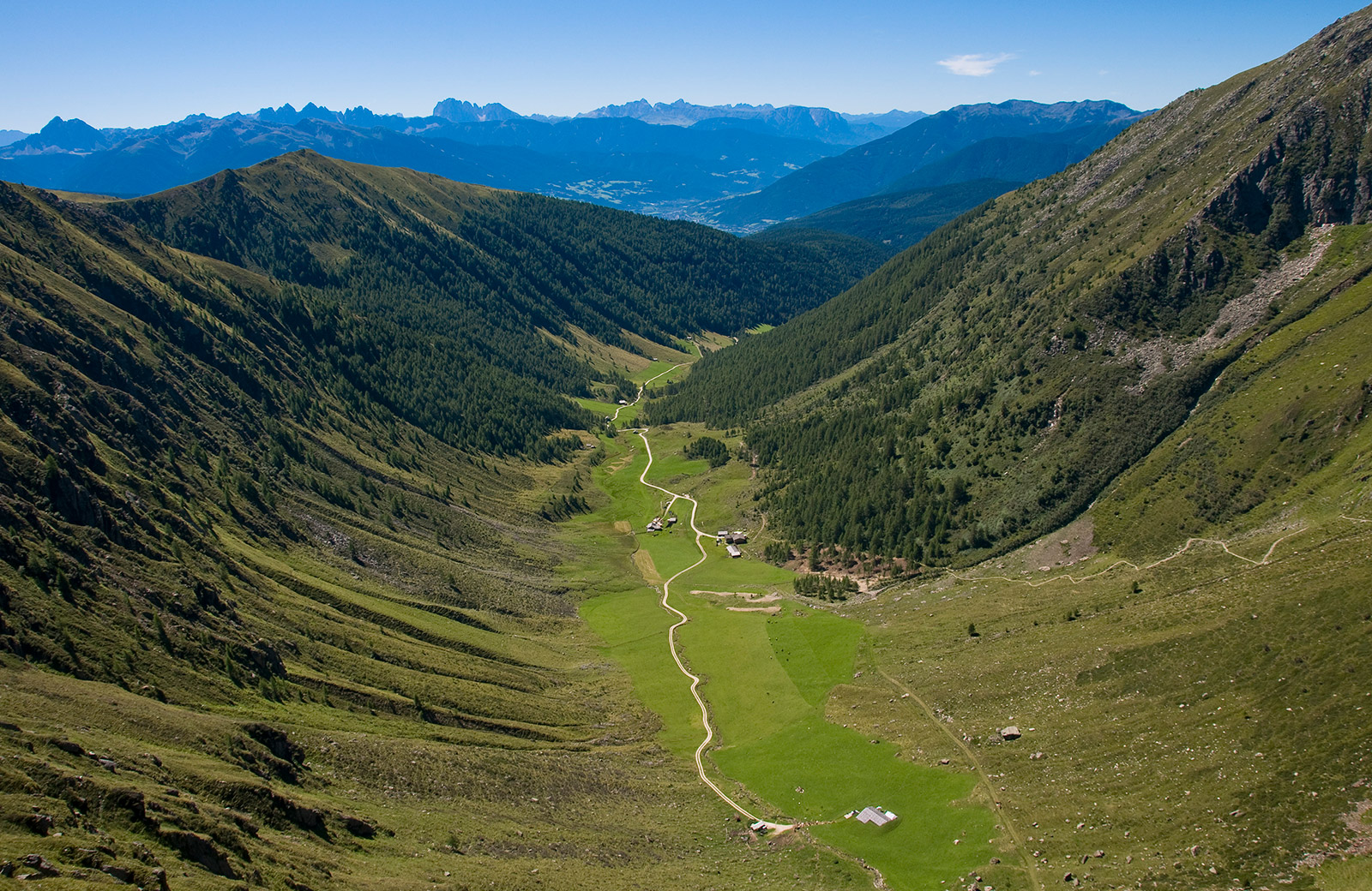 Aerial view of Val Pusteria with the road winding through green pastures and forests, surrounded by the Dolomites.