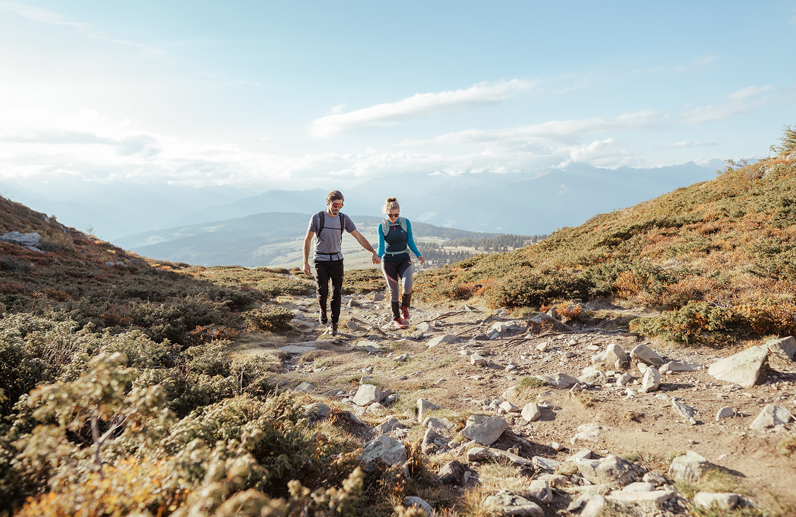 Smiling couple walks hand in hand on a rocky mountain path, with a panoramic view of Val Pusteria.