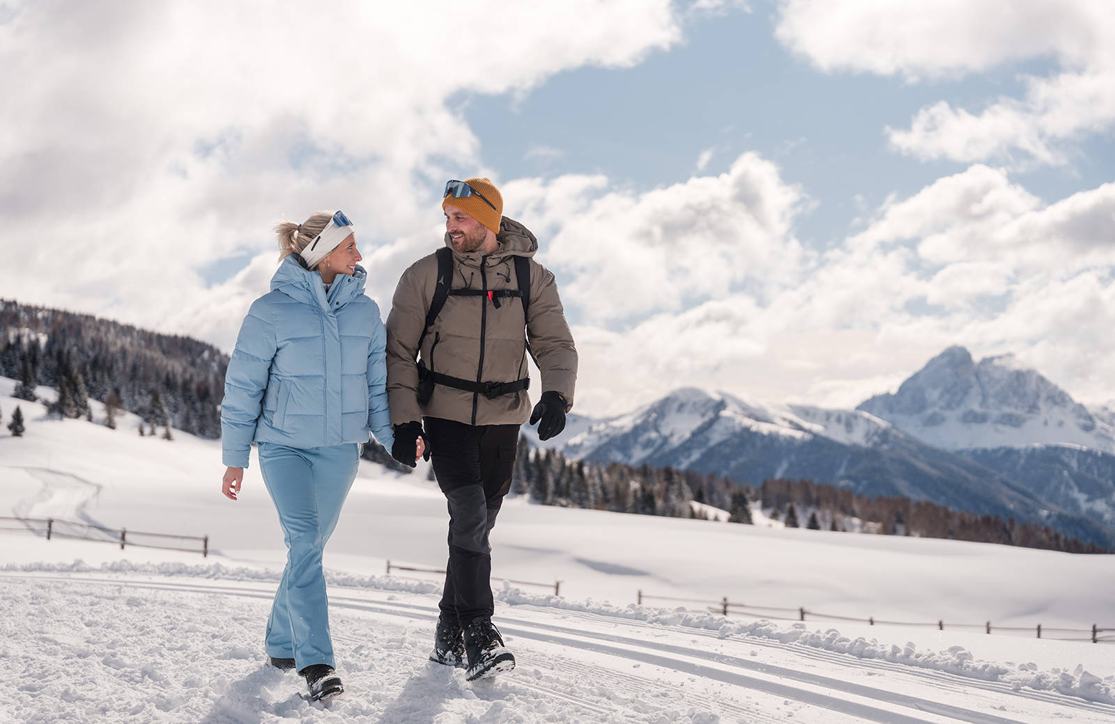 Smiling couple walks on snow, with cross-country skis and Dolomites in the background. Winter holiday in Maranza.
