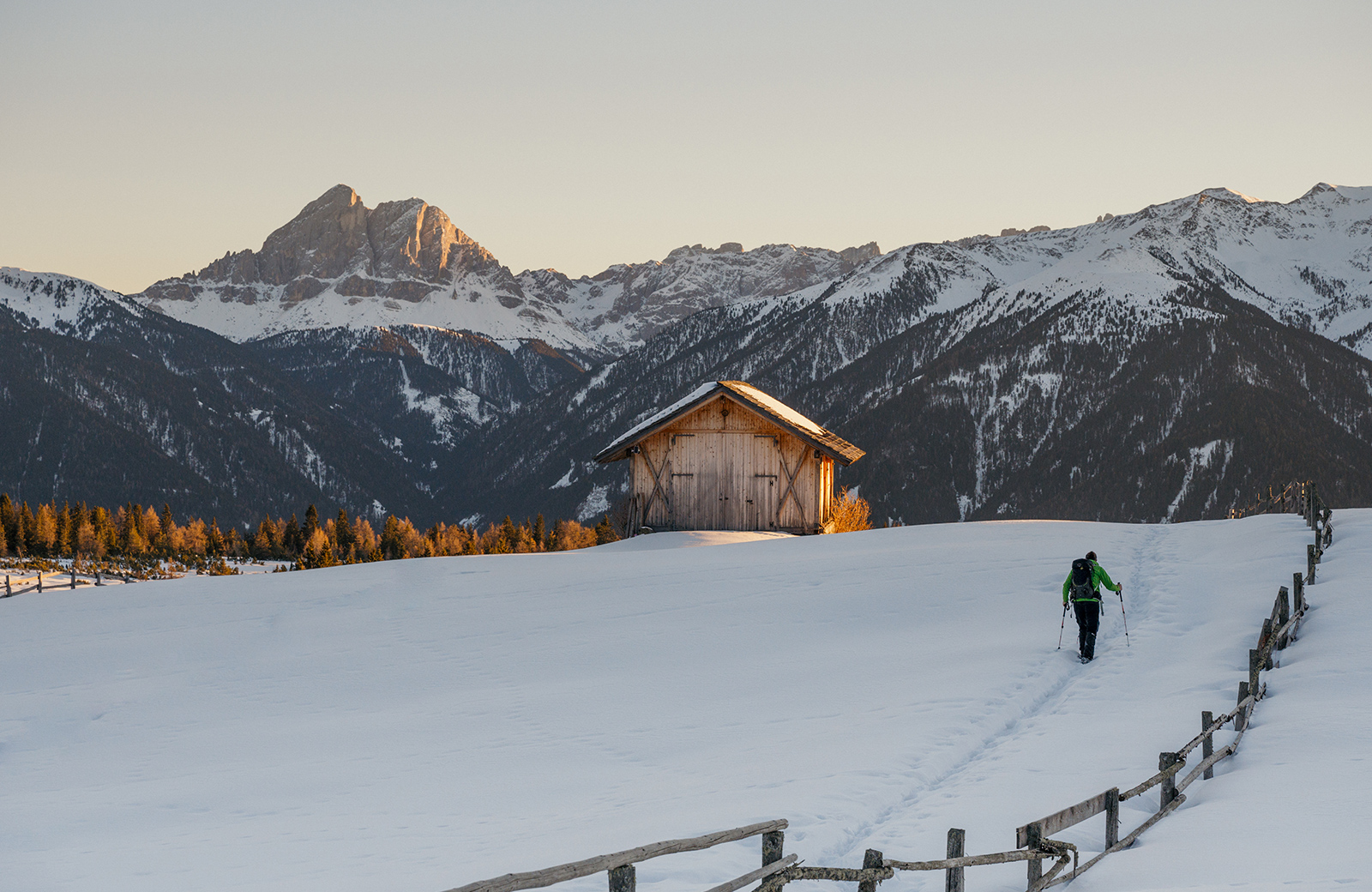 A snowshoer on a snowy path towards a hut, with the Dolomites and Val Pusteria illuminated by the setting sun.