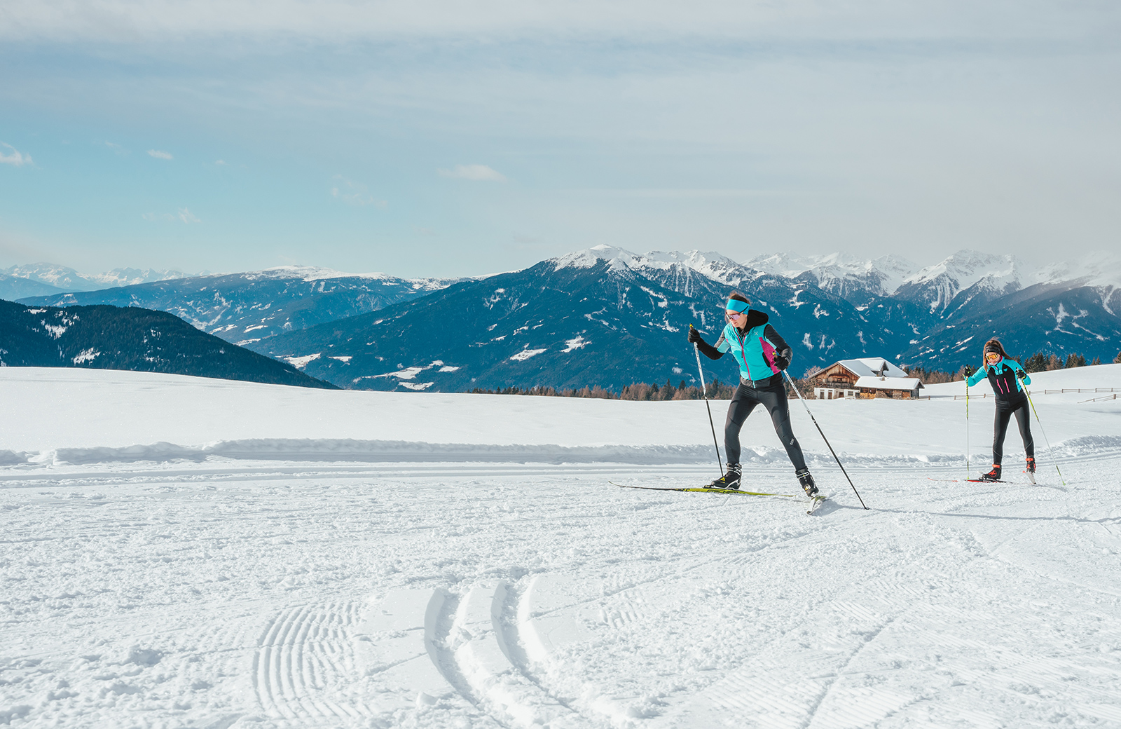 Two people cross-country skiing on a snowy trail in Maranza, with the Dolomites and a mountain hut in the background.