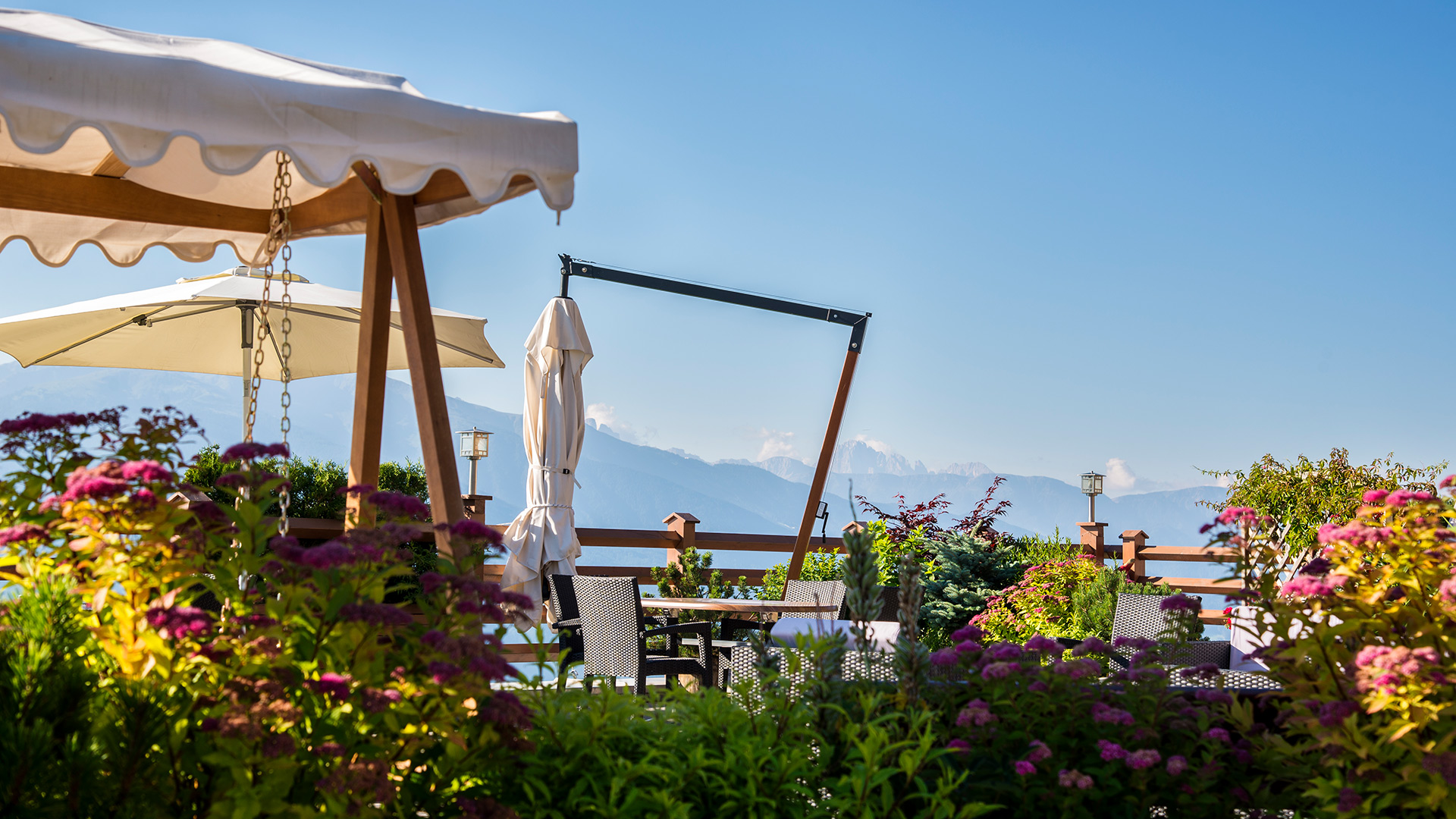 Panoramaterrasse der Pension Wiesenrain mit Tischen und Sonnenschirmen, umgeben von blühendem Grün mit Blick auf die Dolomiten.